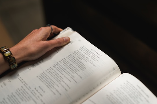 A close-up view of a hand adorned with a bracelet, holding open a book to a page titled 'The Psalms'. The text is printed in a serif font on cream-colored pages, creating a sense of focus and contemplation.