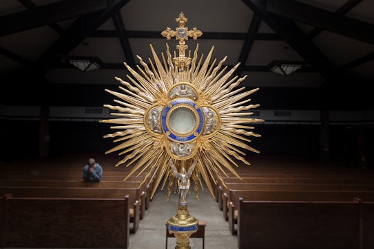 An ornate religious object, a monstrance, featuring a sunburst design with golden metallic rays and intricate detailing is prominently displayed. The background consists of a dimly lit church interior with wooden pews and a vaulted ceiling. There is a lone person sitting in one of the pews, possibly in prayer.