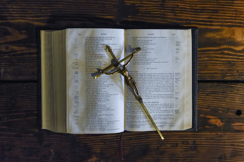 An open Bible displayed on a wooden table with a crucifix placed across its pages. The pages seem to be from the Gospel of John, and the setting suggests a serene and contemplative atmosphere.