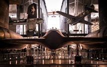 Aviation museum displaying multiple aircraft, including vintage fighter planes, suspended in a spacious and well-lit hangar. The central focus is on a sleek, black aircraft with smooth lines, possibly a stealth or reconnaissance plane. Other aircraft are seen in the background, highlighting the variety of designs from different eras. Visitors are visible below, indicating scale and the public nature of the exhibit.