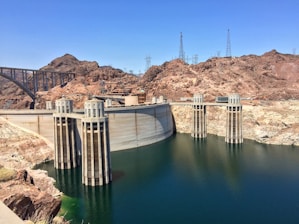 A massive concrete dam with water flowing on one side, surrounded by arid, rocky terrain. The structure features several tall intake towers and power lines are visible against the clear blue sky. There's a large bridge on the left connecting both sides of the canyon.