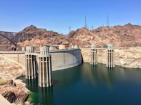 A massive concrete dam with water flowing on one side, surrounded by arid, rocky terrain. The structure features several tall intake towers and power lines are visible against the clear blue sky. There's a large bridge on the left connecting both sides of the canyon.