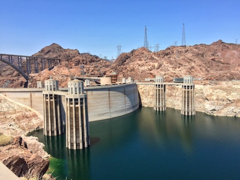 A massive concrete dam with water flowing on one side, surrounded by arid, rocky terrain. The structure features several tall intake towers and power lines are visible against the clear blue sky. There's a large bridge on the left connecting both sides of the canyon.