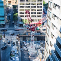 A bustling urban construction site bordered by tall buildings, with a prominent red and white crane extending over the site. Below, construction materials and equipment are organized along the ground. Streets on either side see light traffic with pedestrians and vehicles.