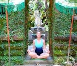 A person is meditating in a garden setting, surrounded by lush green foliage. They are sitting cross-legged on a green mat with eyes closed, wearing a turquoise tank top and dark shorts. Behind them, there is a statue of a deity, and the space is flanked by stone walls covered in vines. Two green parasols with tassels are partially visible at the top corners.