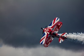 A biplane with a vibrant red, white, and blue color scheme, featuring a Union Jack design on its wings, performs aerobatic maneuvers against a backdrop of dark, stormy clouds. The plane leaves a trail of white smoke as it ascends into the sky.