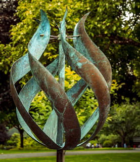 An abstract metal sculpture featuring twisted, elongated elements that resemble flames. The surface has a weathered, greenish-blue patina. The sculpture is set against a lush background of green foliage, indicating a park or garden setting.