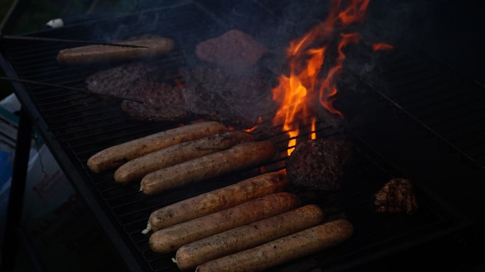 A barbecue grill with several sausages and burger patties cooking over open flames and darkened areas of smoke.