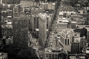A monochrome aerial view of a densely packed urban area featuring a prominent triangular-shaped building at the intersection of two major streets. The architecture varies from modern high-rises to classic stone buildings. The perspective is slightly tilted, offering a dynamic view of the city's layout.