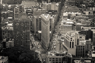 A monochrome aerial view of a densely packed urban area featuring a prominent triangular-shaped building at the intersection of two major streets. The architecture varies from modern high-rises to classic stone buildings. The perspective is slightly tilted, offering a dynamic view of the city's layout.