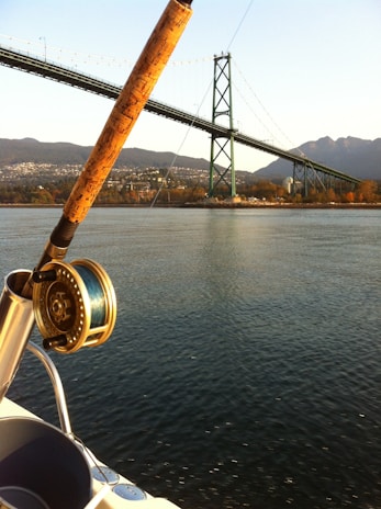 A fishing rod is mounted on a boat, with a scenic view of a large suspension bridge in the background. The water is calm, and the bridge spans across a wide body of water with mountainous terrain in the distance. The fishing reel is prominently displayed in the foreground.