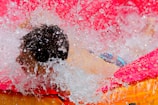 A person is enjoying water splashes in a vivid pink and orange inflatable pool. Droplets of water are captured mid-air, creating a dynamic and joyful scene.