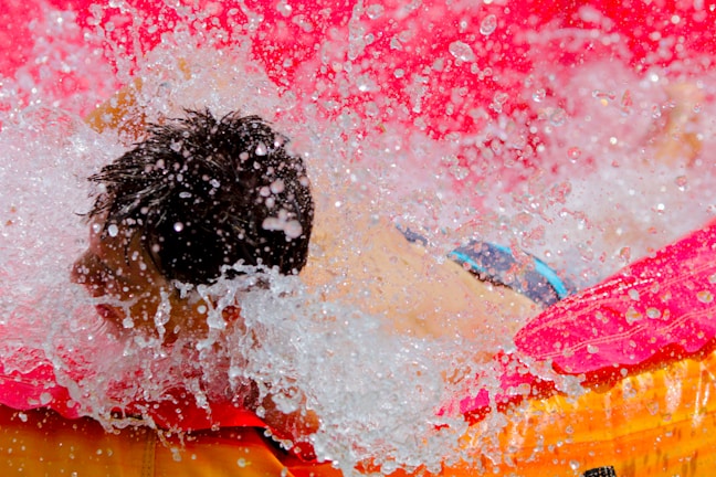 A person is enjoying water splashes in a vivid pink and orange inflatable pool. Droplets of water are captured mid-air, creating a dynamic and joyful scene.