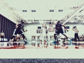 A basketball game is taking place in an indoor court. Players in jerseys are seen in motion, with a player in white attempting to shoot the ball. Referees in striped shirts are visible, and a scoreboard is displayed in the background.