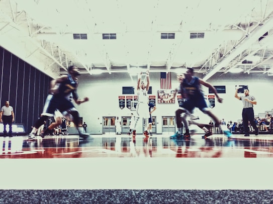 A basketball game is taking place in an indoor court. Players in jerseys are seen in motion, with a player in white attempting to shoot the ball. Referees in striped shirts are visible, and a scoreboard is displayed in the background.