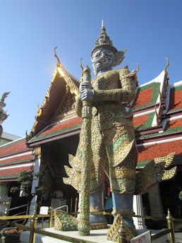 A large, ornate statue of a guardian figure stands in front of a traditional Thai temple. The statue features intricate detailing with vibrant patterns and holds a weapon. The temple behind has a sloped roof with decorative elements and is adorned with red, green, and gold accents.