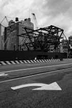 A construction site with large metal framework structures and cranes stands against a backdrop of tall, old buildings. The street in front shows a directional arrow painted on the asphalt, and concrete barriers line the edge of the road. The sky is overcast, adding a dramatic tone to the scene.