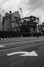 A construction site with large metal framework structures and cranes stands against a backdrop of tall, old buildings. The street in front shows a directional arrow painted on the asphalt, and concrete barriers line the edge of the road. The sky is overcast, adding a dramatic tone to the scene.