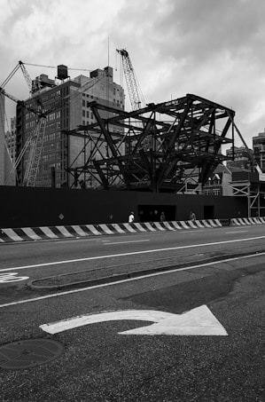 A construction site with large metal framework structures and cranes stands against a backdrop of tall, old buildings. The street in front shows a directional arrow painted on the asphalt, and concrete barriers line the edge of the road. The sky is overcast, adding a dramatic tone to the scene.