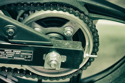 A close-up view of a bicycle or motorcycle drive chain and gear mechanism. The chain wraps around a metal sprocket, which is secured with nuts and bolts. A part of the frame can be seen, along with a label that provides information about the chain's tension and lubrication.