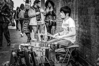 A black and white photograph of a street musician playing a drum set. The musician, wearing a T-shirt and jeans, is engrossed in his performance. A group of people stand nearby, some in winter coats, seemingly observing the performance while walking along a cobblestone street next to a brick wall.