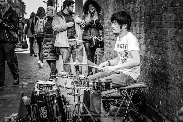 A black and white photograph of a street musician playing a drum set. The musician, wearing a T-shirt and jeans, is engrossed in his performance. A group of people stand nearby, some in winter coats, seemingly observing the performance while walking along a cobblestone street next to a brick wall.