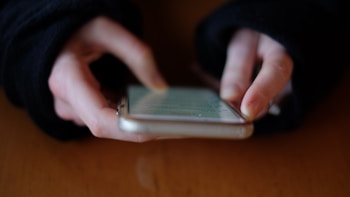 Hands are holding a smartphone, with fingers actively interacting with the touch screen. The background is out of focus, drawing attention to the device and the hands.