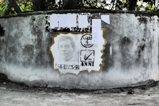A weathered concrete wall displays a stencil portrait, surrounded by election-themed graphics. Above the portrait, several papers are attached to the wall, likely serving as notices or posters.