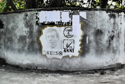 A weathered concrete wall displays a stencil portrait, surrounded by election-themed graphics. Above the portrait, several papers are attached to the wall, likely serving as notices or posters.