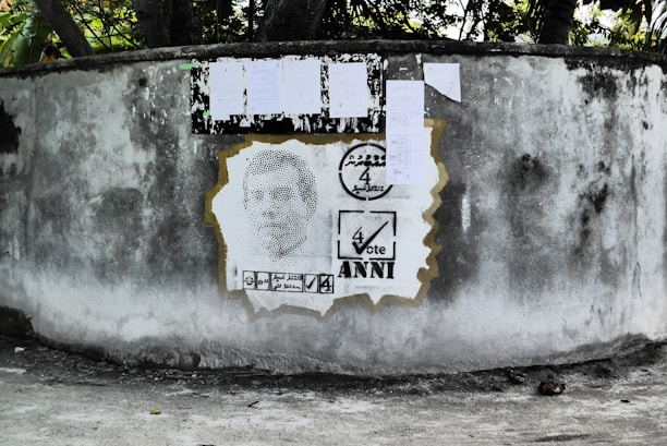 A weathered concrete wall displays a stencil portrait, surrounded by election-themed graphics. Above the portrait, several papers are attached to the wall, likely serving as notices or posters.