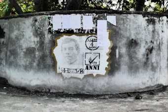 A weathered concrete wall displays a stencil portrait, surrounded by election-themed graphics. Above the portrait, several papers are attached to the wall, likely serving as notices or posters.