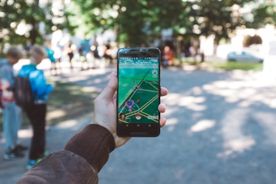 A person is holding a smartphone with a gaming app open, featuring an augmented reality map. The setting is outdoors, with people visible in the background, suggesting a park or public space.