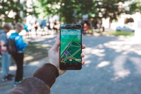 A person is holding a smartphone with a gaming app open, featuring an augmented reality map. The setting is outdoors, with people visible in the background, suggesting a park or public space.