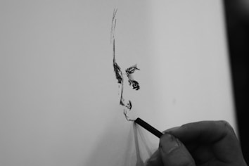 A hand is holding a drawing tool, sketching the outline of a face on a blank sheet of paper. The artwork appears to be in its early stages with minimal detail, focusing on the facial features.