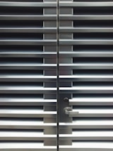 A metallic or aluminum louvered door with horizontal slats and a silver handle on one side. The door is symmetrical and provides a sleek, industrial aesthetic.