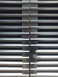 A metallic or aluminum louvered door with horizontal slats and a silver handle on one side. The door is symmetrical and provides a sleek, industrial aesthetic.