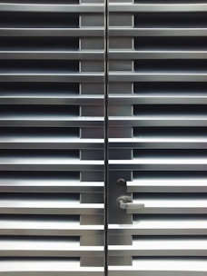 A metallic or aluminum louvered door with horizontal slats and a silver handle on one side. The door is symmetrical and provides a sleek, industrial aesthetic.