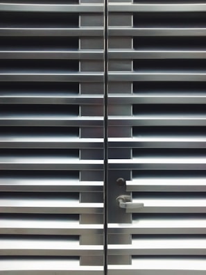 A metallic or aluminum louvered door with horizontal slats and a silver handle on one side. The door is symmetrical and provides a sleek, industrial aesthetic.
