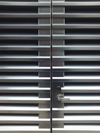 A metallic or aluminum louvered door with horizontal slats and a silver handle on one side. The door is symmetrical and provides a sleek, industrial aesthetic.