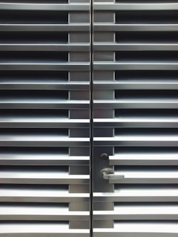 A metallic or aluminum louvered door with horizontal slats and a silver handle on one side. The door is symmetrical and provides a sleek, industrial aesthetic.