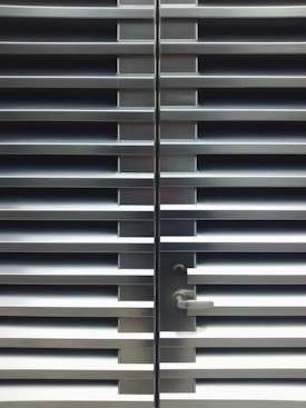 A metallic or aluminum louvered door with horizontal slats and a silver handle on one side. The door is symmetrical and provides a sleek, industrial aesthetic.
