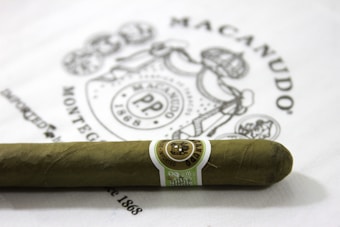 A cigar with a greenish wrapper and a band wrapped around it featuring intricate designs and text. The background includes a partially visible printed logo and text, adding to the brand's aesthetic.