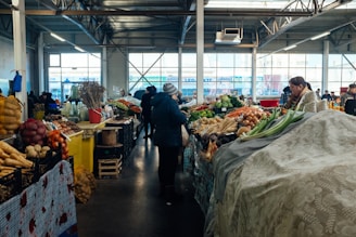 A bustling indoor market scene with vendors and customers engaged around stalls displaying a variety of fresh produce. Baskets and containers are filled with vegetables like carrots, potatoes, and onions, while other items such as bottled goods and dry herbs are also visible. Natural light pours in through large windows, illuminating the industrial-style space.