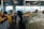 A bustling indoor market scene with vendors and customers engaged around stalls displaying a variety of fresh produce. Baskets and containers are filled with vegetables like carrots, potatoes, and onions, while other items such as bottled goods and dry herbs are also visible. Natural light pours in through large windows, illuminating the industrial-style space.