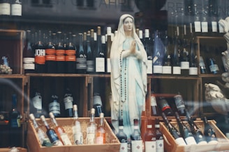 A display features a statue of a religious figure, adorned with a rosary, amidst a collection of various wine bottles. The bottles are organized on wooden crates, with some positioned upright and others lying flat. The background includes additional bottles and elements of the store's decor.