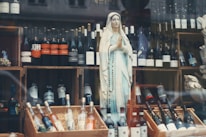A display features a statue of a religious figure, adorned with a rosary, amidst a collection of various wine bottles. The bottles are organized on wooden crates, with some positioned upright and others lying flat. The background includes additional bottles and elements of the store's decor.