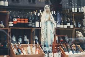 A display features a statue of a religious figure, adorned with a rosary, amidst a collection of various wine bottles. The bottles are organized on wooden crates, with some positioned upright and others lying flat. The background includes additional bottles and elements of the store's decor.