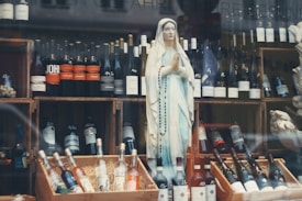 A display features a statue of a religious figure, adorned with a rosary, amidst a collection of various wine bottles. The bottles are organized on wooden crates, with some positioned upright and others lying flat. The background includes additional bottles and elements of the store's decor.