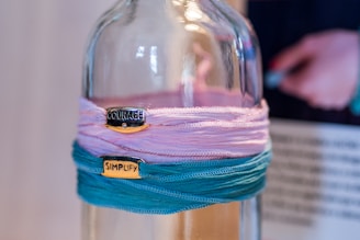 A glass bottle is adorned with colorful fabric wraps in shades of pink and teal. Two metallic tags with words 'COURAGE' and 'SIMPLIFY' are attached to the fabrics. In the background, a blurred figure appears holding something.