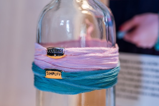A glass bottle is adorned with colorful fabric wraps in shades of pink and teal. Two metallic tags with words 'COURAGE' and 'SIMPLIFY' are attached to the fabrics. In the background, a blurred figure appears holding something.